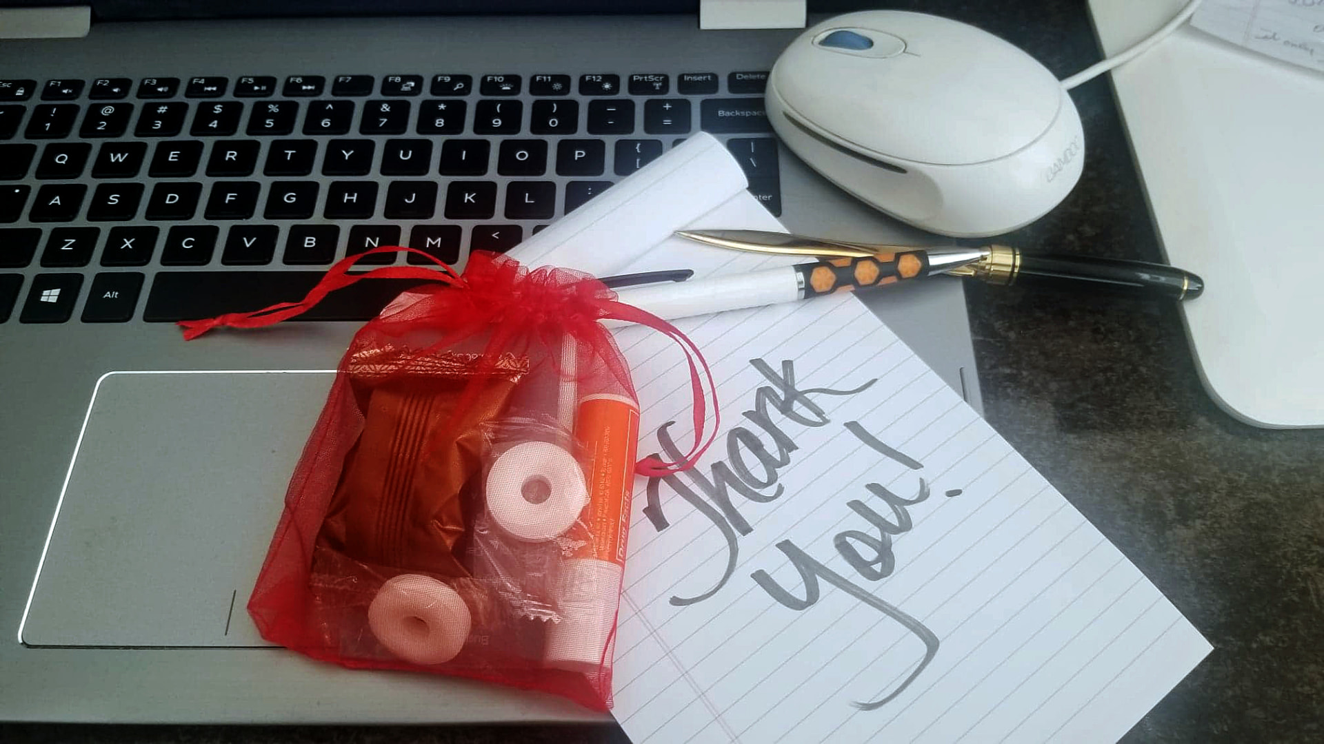 A handwritten thank-you note and a small red gift bag of treats resting on a laptop keyboard beside a pen and mouse