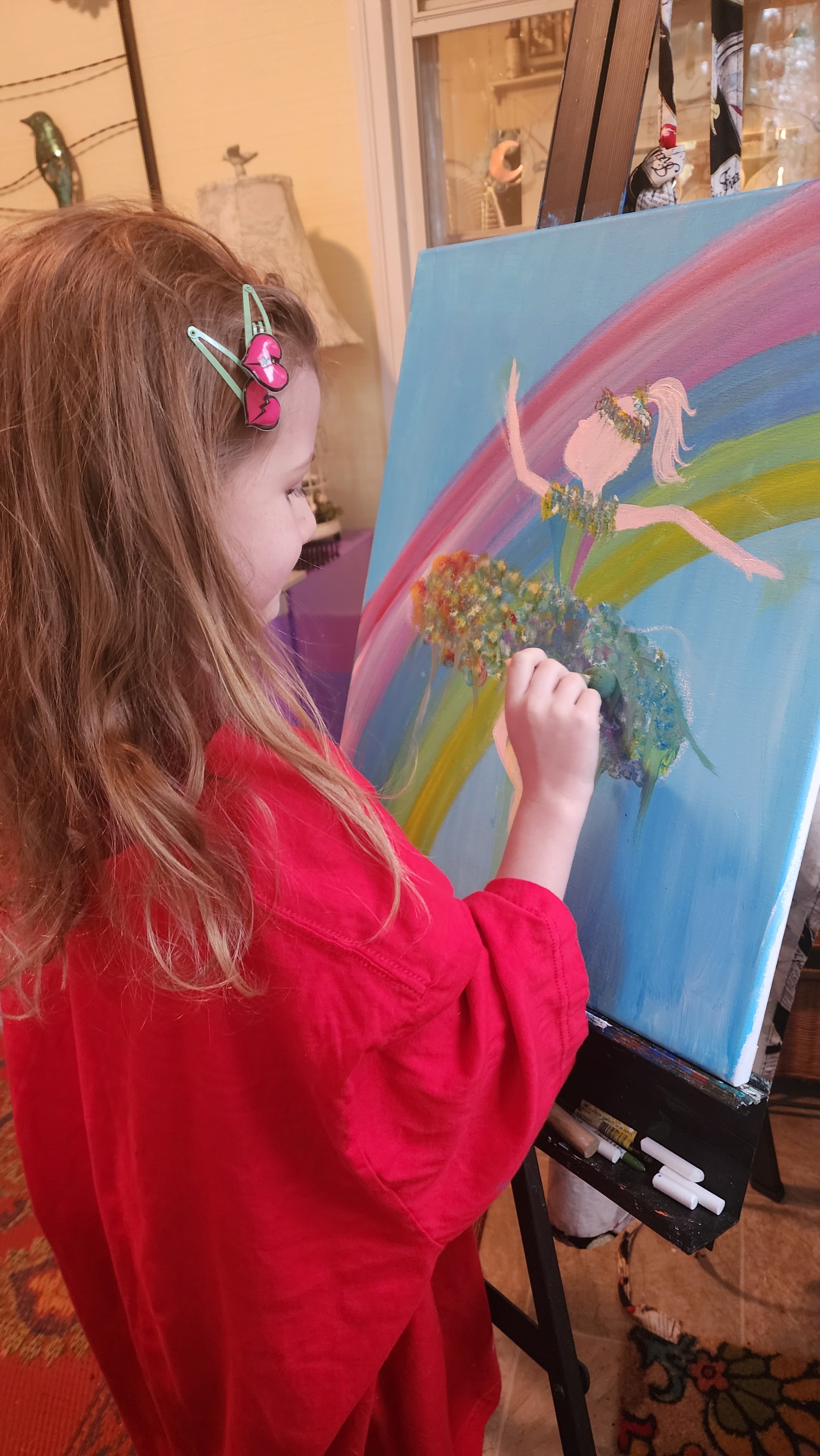 A young girl painting a fairy and rainbow scene on a canvas at an easel