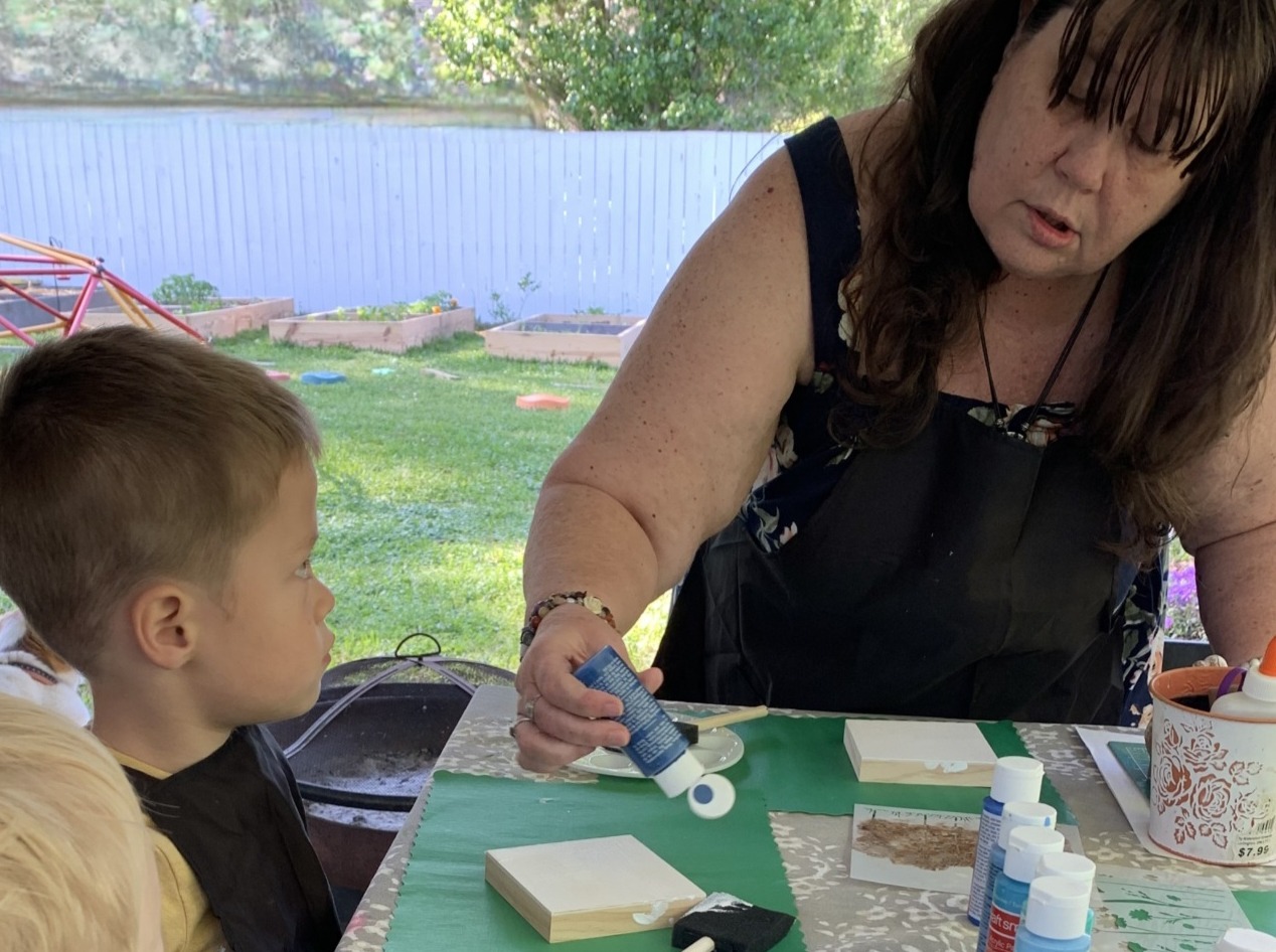 Michelle squeezing blue paint onto a small wooden block while a young boy watches at an outdoor craft table